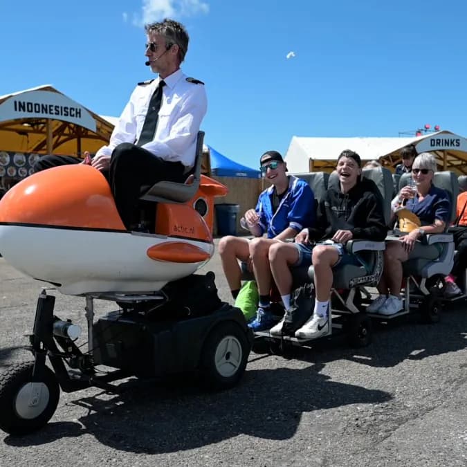 Een piloot met headset zit op een oranje mini-vliegtuigje op wielen en trekt een sliert vliegtuigstoelen met lachende passagiers over het plein.