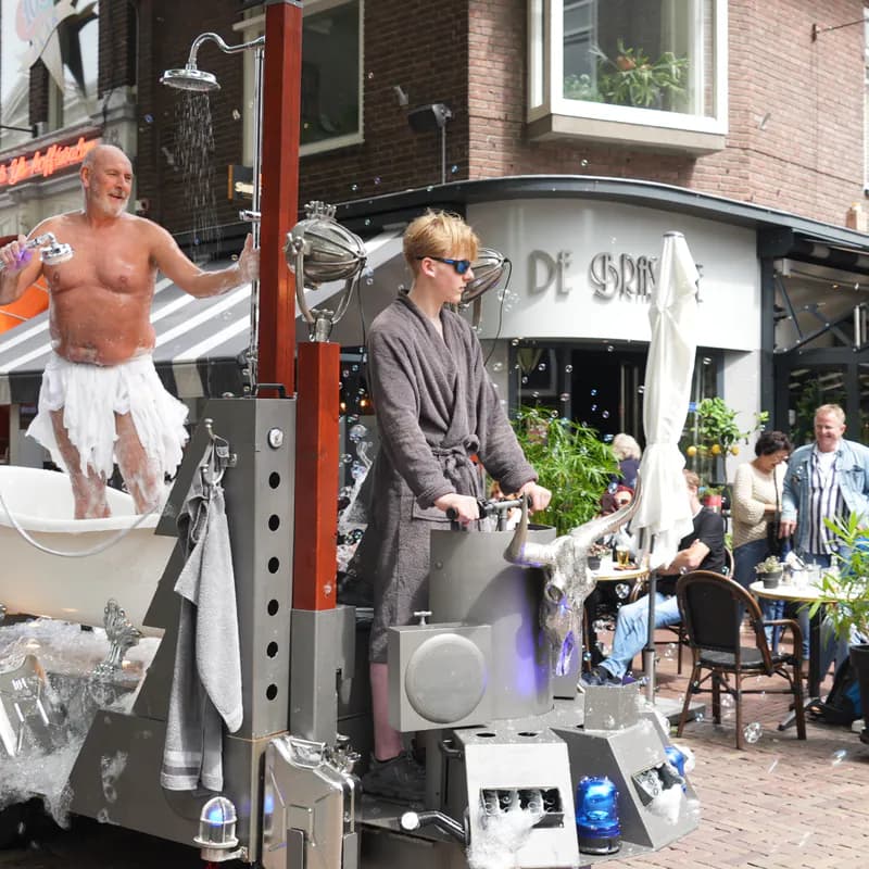 Het bad staat op een metalen kar met een douchekop erboven, en een tweede speler in een grijze badjas bedient de kar terwijl die langs een terras rijdt.