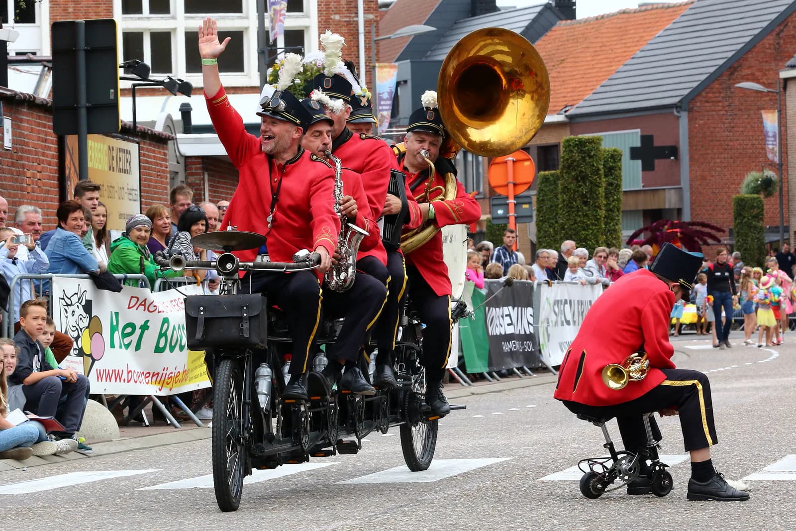 Een fanfare in rode uniformjassen fietst op een lange tandem door de straat, voorop knielt een muzikant op een klein wieltjeswagentje en speelt trompet, terwijl een van zijn collega's naar het publiek zwaait.