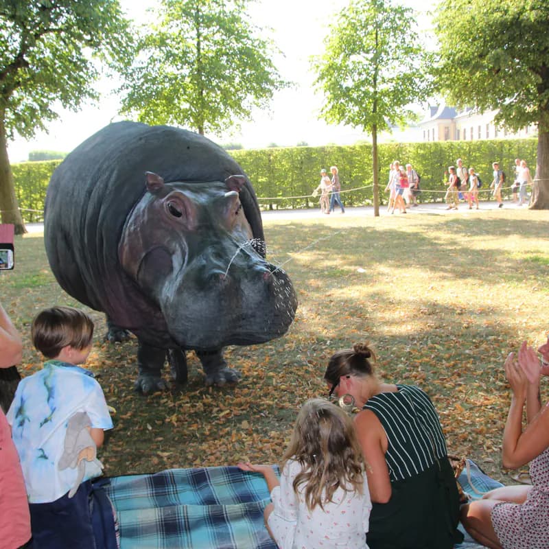 Het nijlpaard staat op het gras in een park en spuit water uit haar neus, terwijl kinderen op een picknickkleed toekijken.