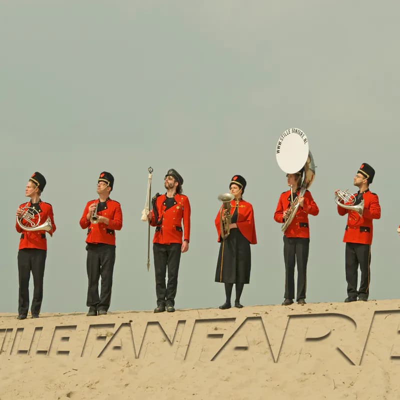 Zes muzikanten in rode jassen staan op een rij op het strand, met voor hen de letters van DE STILLE FANFARE uitgesneden in het zand, aan beide zijden doorlopend tot buiten beeld.