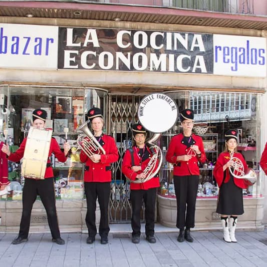 Zeven muzikanten van De Stille Fanfare in rode uniformjassen met zwarte petten staan op een rij voor een winkelpui, met trom, sousafoon, hoorns en een vaandeldrager.