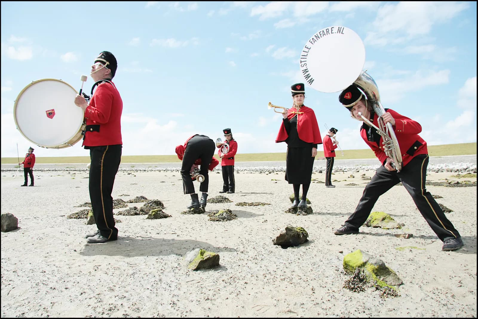 Zeven muzikanten in rode uniformjassen met zwarte petten spelen op een grote trom, een sousafoon en trompetten, verspreid over een open vlakte onder een blauwe lucht.